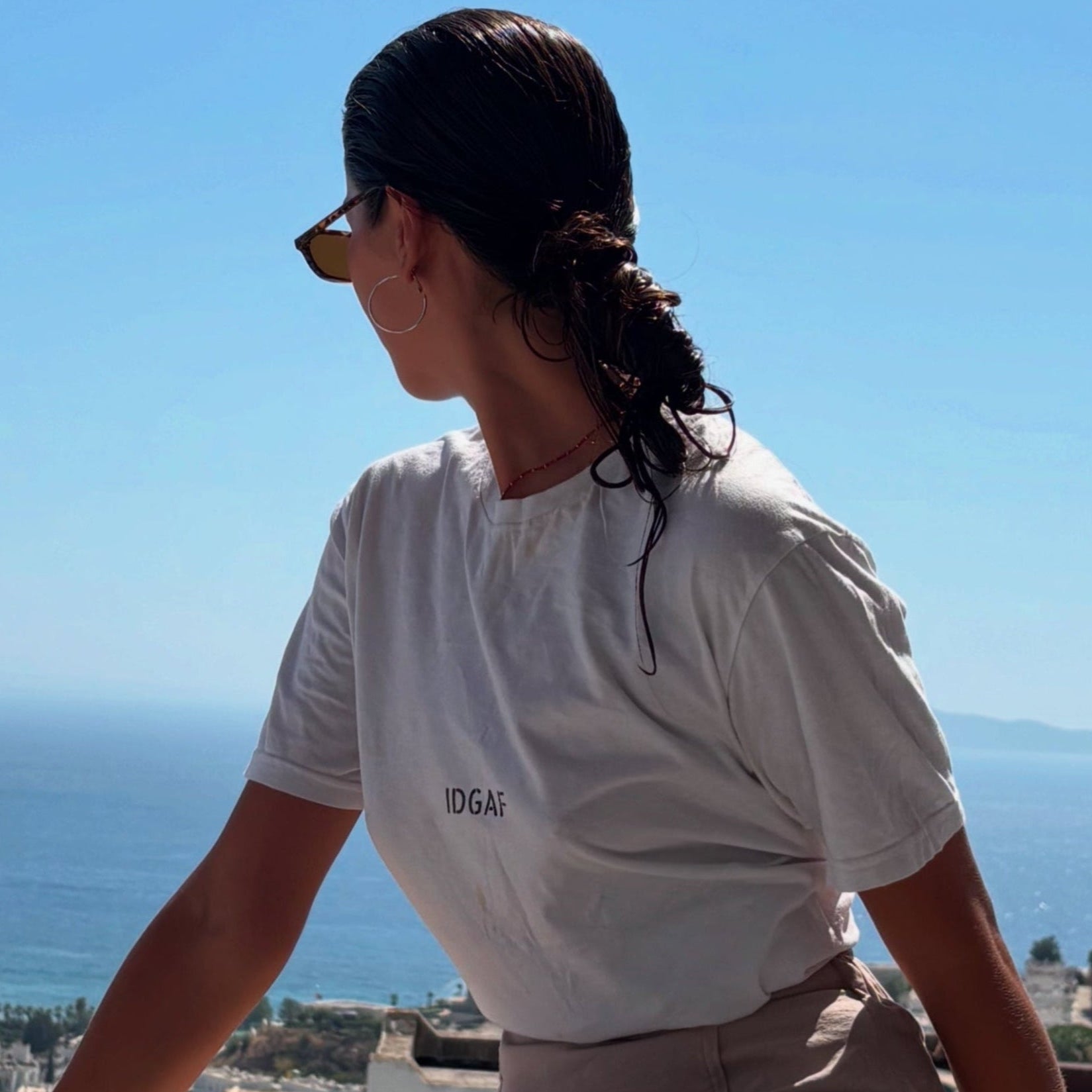 Person wearing a white t-shirt with text, sitting on a ledge overlooking a scenic view of the ocean and buildings.